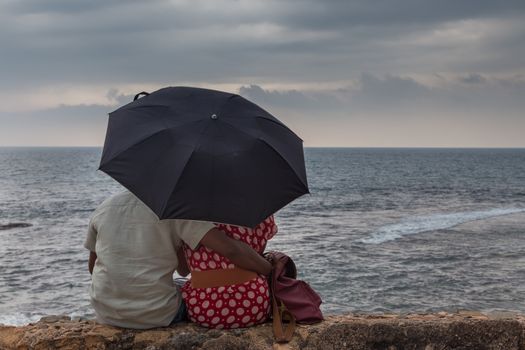 Rear View of a Indian couple sitting by sea with an umbrella looking at storm on ocean horizon.