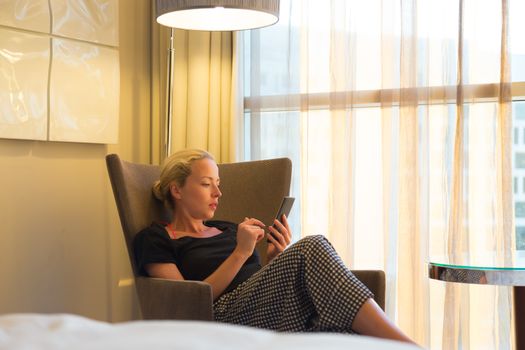 Casually dressed attractive blonde caucasian woman holding and using smart phone while sitting on armchair in modern hotel room.