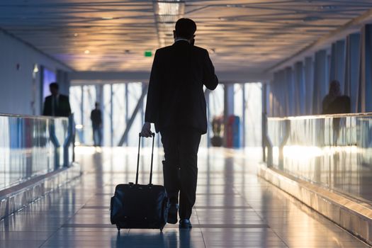 Businessman dragging a small carry on luggage suitcase at airport corridor walking to departure gates.