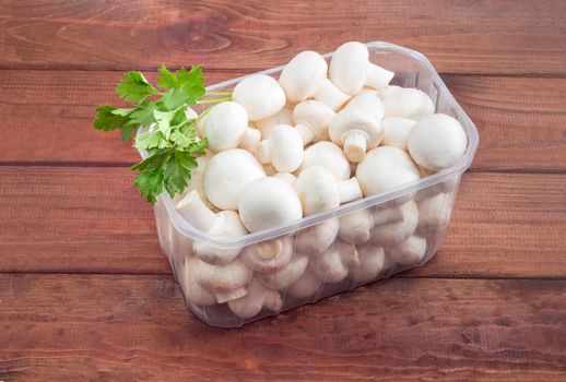 Fresh uncooked button mushrooms in a transparent plastic tray and sprig of parsley on a dark wooden surface
