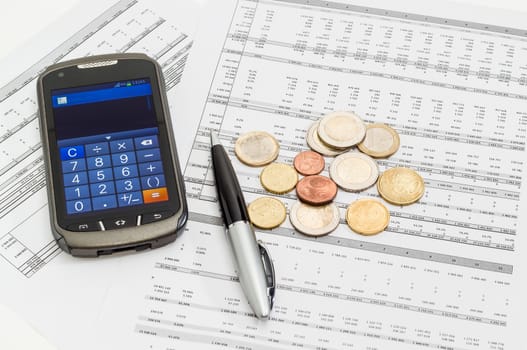 Several euro coins different denominations on the printed table with data, pen and modern smartphone in calculator mode 
