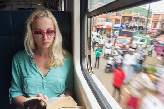 Blonde caucasian woman reading book by train window , traveling by train in third world country. Blured people on street can be seen trough window.