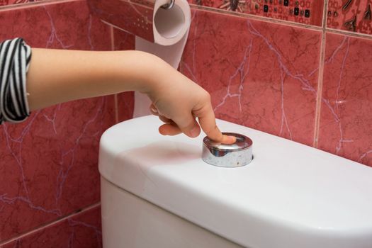 Child's hand finger presses the button on the water draining into the toilet bowl, close-up