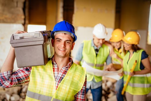 Smiling young electrician with helmet holding toolbox at a construction site and looking at camera. His architect colleagues review plan in background.