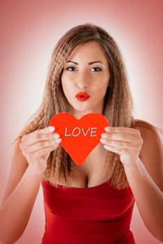 Beautiful smiling girl holding a red heart that says Love and sending a kiss. Looking at camera.