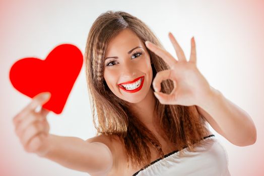 Close-up of a beautiful smiling girl holding a red heart and pointing sign ok. Selective focus. Focus on the girl. Looking at camera.