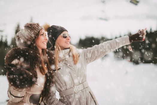 Two beautiful happy female friends enjoying in winter vacations. They taking a walk while its snowing and looking away.