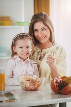 Mother and daughter having breakfast in the kitchen. They eating fruit salad. 