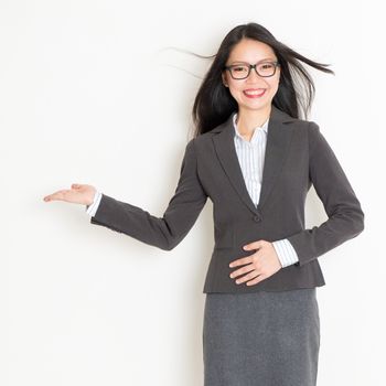 Portrait of Asian businesswoman in formalwear smiling and hand showing something, standing on plain background.
