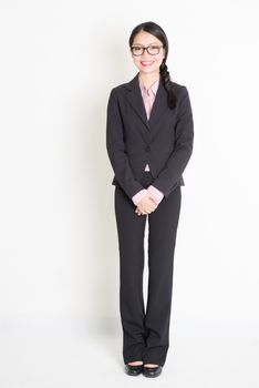 Full body portrait of young Asian businesswoman in formalwear smiling, standing on plain background.