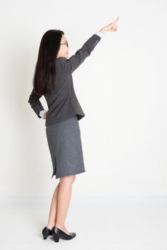 Rear view of Asian businesswoman in formalwear hand pointing away and smiling, full body standing on plain background.