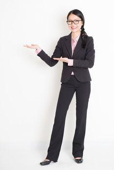 Full body portrait of young Asian businesswoman in formalwear hands showing something and smiling, standing on plain background.