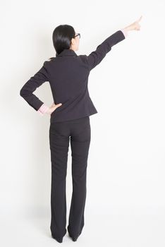 Full body back view of young Asian businesswoman in formalwear finger pointing on something, standing on plain background.