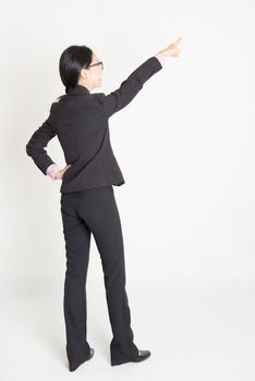 Full body rear view of young Asian businesswoman in formalwear finger pointing on something, standing on plain background.