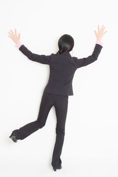 Full body back view of young Asian businesswoman in formalwear banging and leaning on wall, standing on plain background.