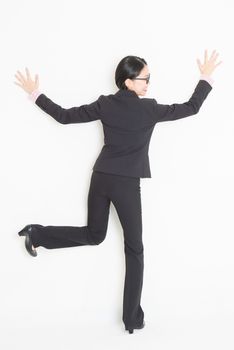 Full length back view of young Asian businesswoman in formalwear hands and body leaning on wall, standing on plain background.