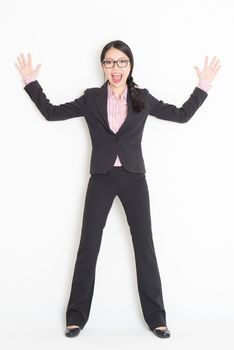 Full length front view of shocked young Asian businesswoman in formalwear leaning on wall, standing on plain background.