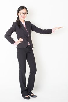 Full length front view of young Asian businesswoman in formalwear hand displaying something, standing on plain background.