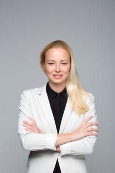 Portrait of cheerful, beautiful, smart, young businesswoman in white business attire, standing with arms crossed against gray background. Copy space on top.