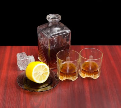 Decanter and two glasses with whiskey, stopper from the decanter and lemon on a saucer on a wooden table on a dark background
