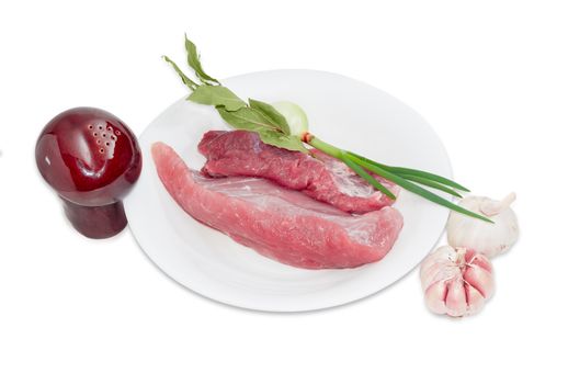 Several pieces of a fresh uncooked pork, branch of dried bay leaves, green onion on white plate, pepper shaker and two garlic bulbs on a light background 
