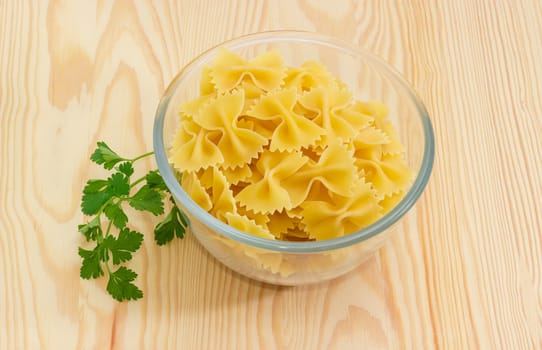 Uncooked bow-tie pasta in glass bowl and sprig of a parsley on a light wooden surface
