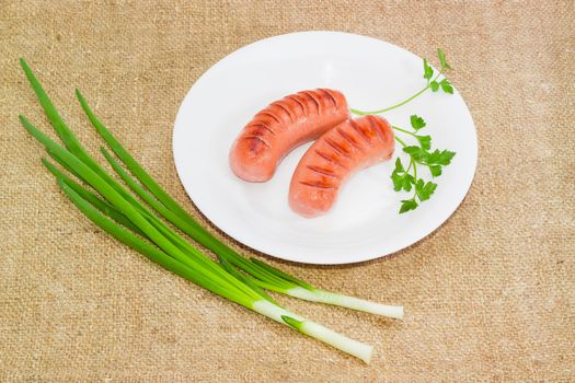 Two grilled thick frankfurters and sprig of parsley on a white dish, green onion on a sackcloth
