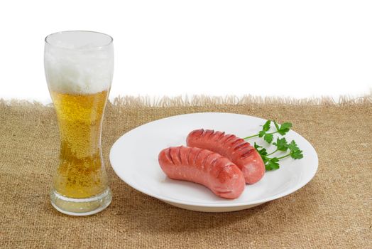 Two fried thick frankfurters and a sprig of parsley on a white dish and glass of a lager beer on a surface by sackcloth on a light background
