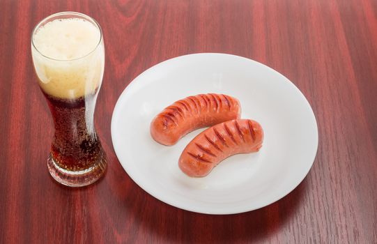 Two fried thick frankfurters on a white dish and glass of a dark beer on a wooden table
