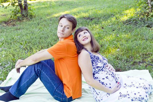 Husband and pregnant wife sitting on green grass in summer time
