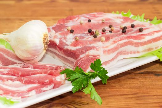 Uncooked partly sliced streaky pork belly bacon with spice, garlic and greens on a white dish closeup on a old wooden surface
