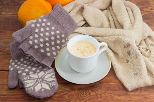 White cup of coffee with cream on a background of women's woolen hybrid mittens and knitted scarf and oranges on wooden surface
