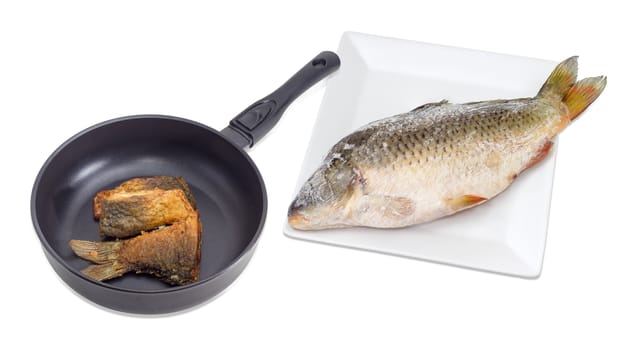 Several fried slices of carp on an aluminium cast frying pan with ceramic non-stick coating and whole frozen carp on a white square dish beside on a light background
