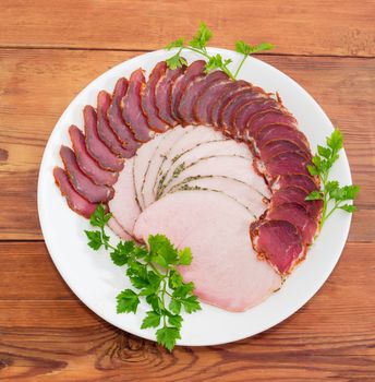 Sliced dried pork tenderloin and ham with twigs of parsley on a white dish on a wooden surface
