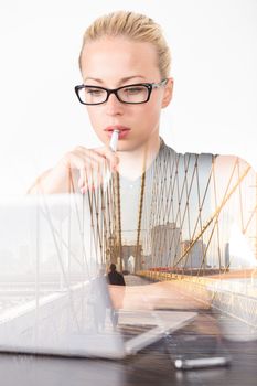 Business woman wearing black glasses, holding pen in hand, checking some information on her laptop computer in office, of thougths, day dreaming about afterwork leisure activities in New York city.