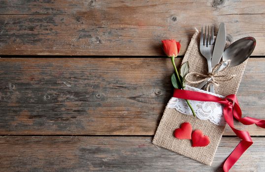 Cutlery set inside pouch with hearts, red rose and silk ribbon over a wooden background