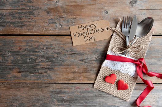 Valentines day label attached to cutlery inside a pouch with decorative ribbon over a wooden background