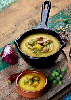 Arrangement of Pea Soup with Smoked Sausages in Black Iron Cast and Clay Bowl with Raw Ingredients and Wooden Spoon closeup on Cracked Wooden background