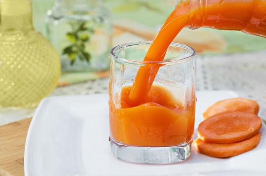 Carrot juice is poured from the bottle into the glass, which stands on the table and a white plate. Selective focus.