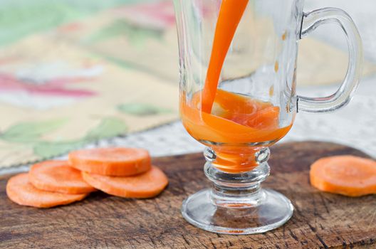 Fresh carrot juice is poured into glass Cup on a stem, which stands on the table and chopping Board, close can not see the edge. Slices of carrots lying around. Selective focus.