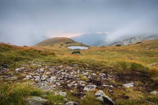 Foggy Mountain Landscape with a Tarn and Rocks in Foreground at Sunset