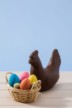 Easter chocolate chicken and colored eggs in a small basket over a white table with blue background