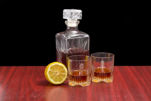Decanter and two glasses with whiskey and lemon cut in half on a wooden table on a dark background
