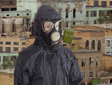 Person in a rubber gas mask and in a black jacket with a hood against the backdrop of abandoned and partially destroyed industrial buildings
