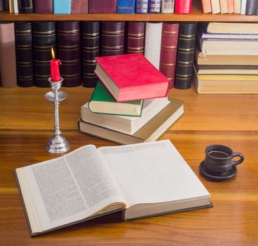 Open big book, stack of books different formats and cover design, cup of coffee and a burning candle in candlestick on a wooden table against the background of shelf with books
