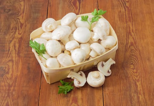 Fresh cultivated button mushrooms and twigs of parsley in the wooden basket, one whole mushroom and mushroom cut in half separately beside on an old wooden surface
