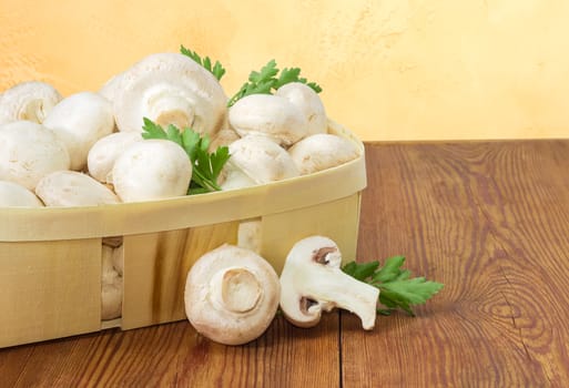 Fragment of wooden basket with fresh cultivated button mushrooms and twigs of parsley, one whole mushroom and one half of mushroom separately beside on an old wooden surface
