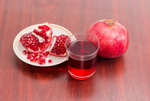 Pomegranate juice in a glass, one ripe whole pomegranate, part of the fresh split pomegranate on a saucer on a red wooden surface
