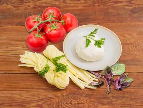 Piece of the fresh mozzarella cheese on the saucer, two portion of the mozzarella cheese, twisted to form a plait, cluster of tomatoes and twigs of parsley and purple basil on an old wooden surface
