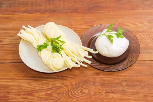 Two portion of the mozzarella cheese in the shape of strings, twisted to form a plait and ball of the fresh mozzarella cheese on the glass saucer with twigs of parsley on an old wooden surface
